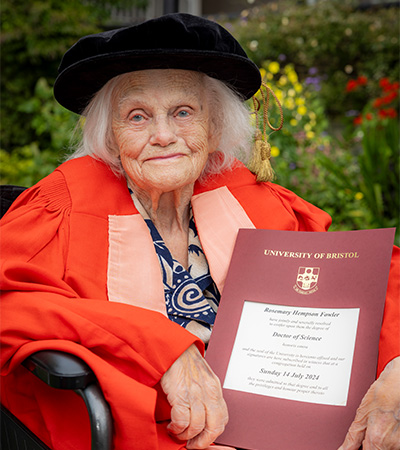 An image of Rosemary Fowler at Darwin College in Cambridge wearing a red Honorary Degree gown smiling and holding her degree